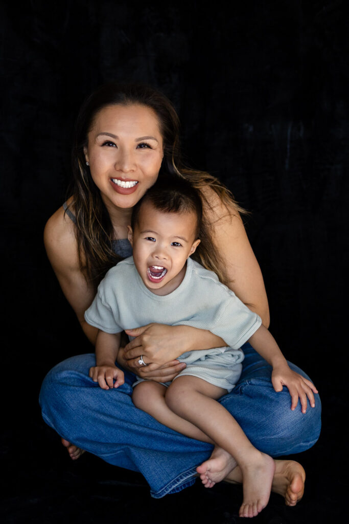 A mother sits cross legged holding her toddler as he laughs openly toward the camera during Motherhood Photo Sessions