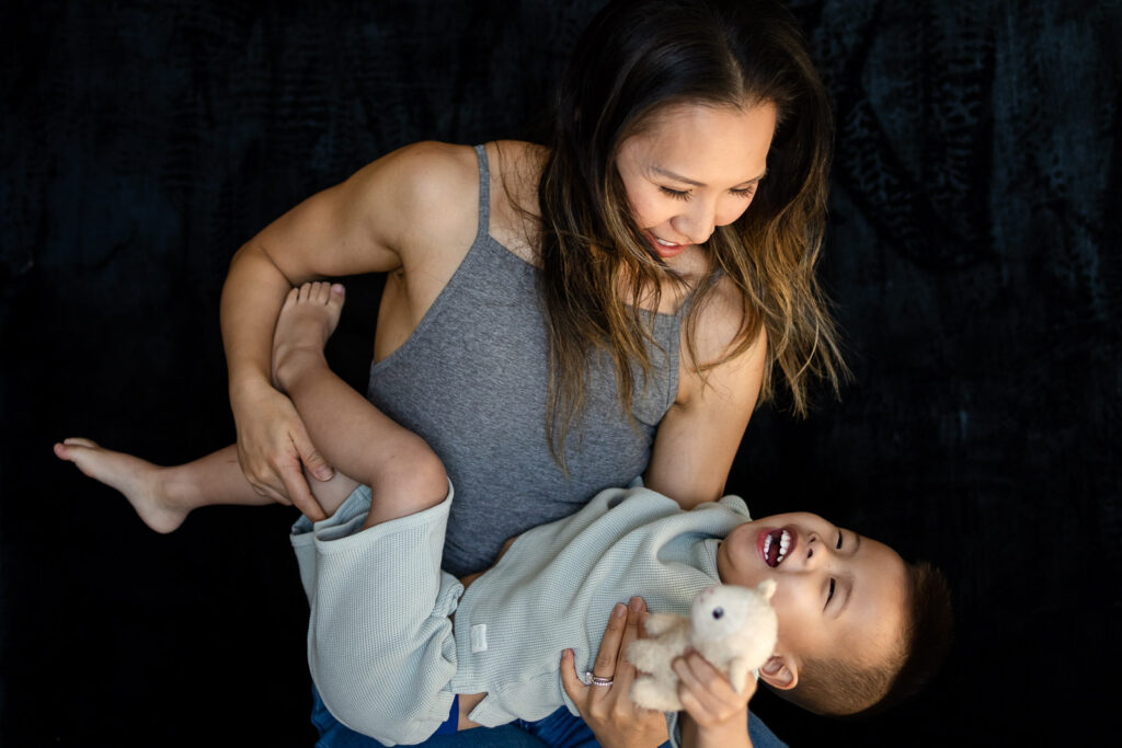 A mother laughs while playfully holding her toddler upside down as he giggles and clutches a small stuffed animal during Mom and Me mini sessions