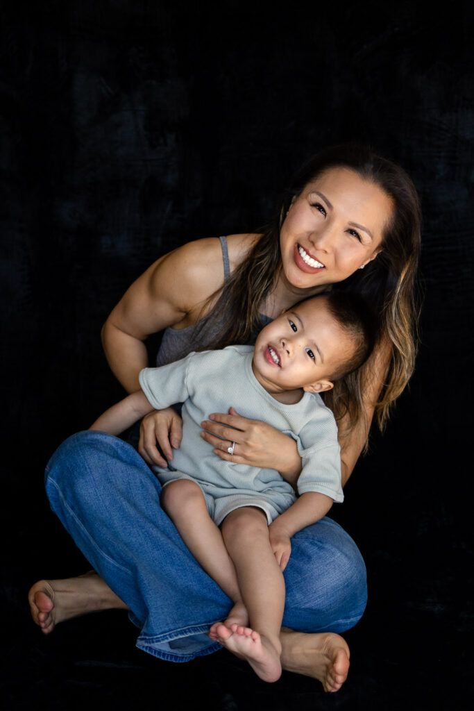A mother sits cross legged holding her toddler close as both smile gently toward the camera in a quiet studio moment.