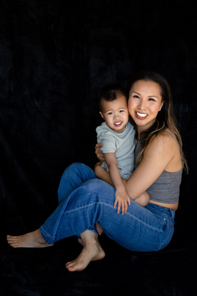 A mother sits on the floor holding her toddler close as they both look toward the camera with relaxed happy expressions during Motherhood Photo Sessions