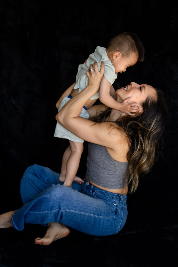 A mother lifts her toddler up toward her face as they laugh and touch cheeks during a playful moment during Mama & Me Photos
