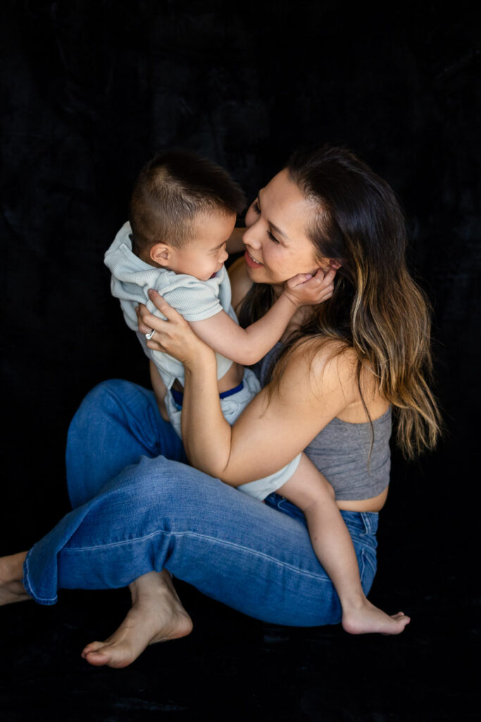 A mother and toddler sit facing each other as he reaches up to her face and she smiles softly back at him during Mama & Me Photos