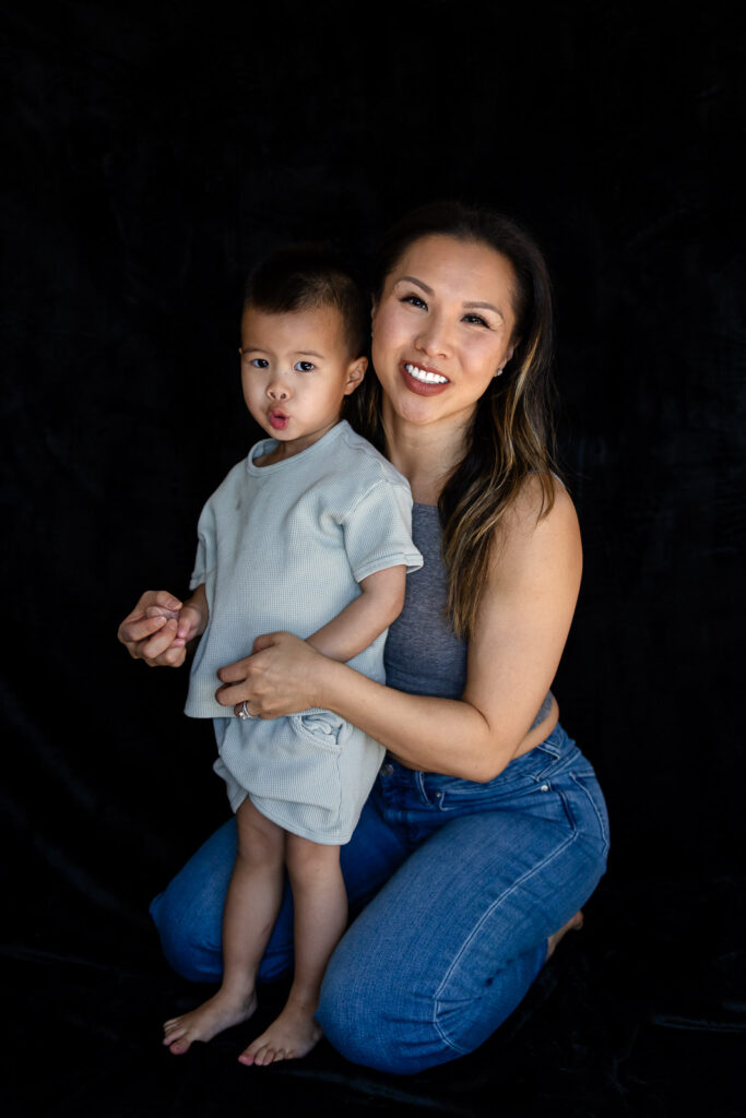 A mother kneels behind her toddler, smiling at the camera as he makes a playful expression during Motherhood Mini Sessions.