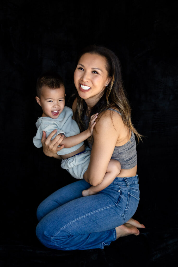 A mother kneels holding her toddler on her hip as both smile toward the camera in a relaxed pose.