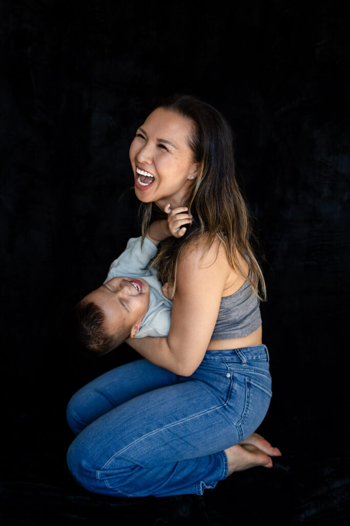A mother laughs openly as her toddler leans back in her arms during a joyful playful moment.
