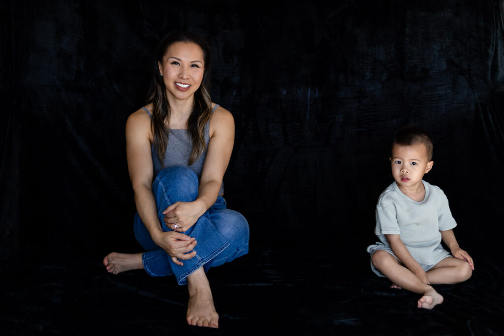 A mother sits on the floor with her toddler nearby as both look toward the camera in a calm candid moment during Motherhood Mini Sessions.