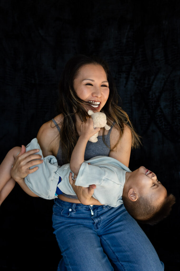 A mother laughs with her eyes closed as her toddler rests across her arms, sharing a joyful playful moment during Mom and Me mini sessions