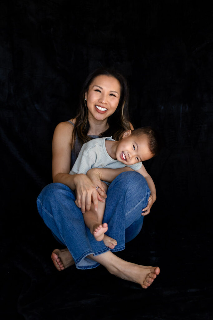 A mother sits cross legged on the studio floor smiling warmly as her toddler rests his head on her knees and grins toward the camera for a Motherhood Mini Sessions.