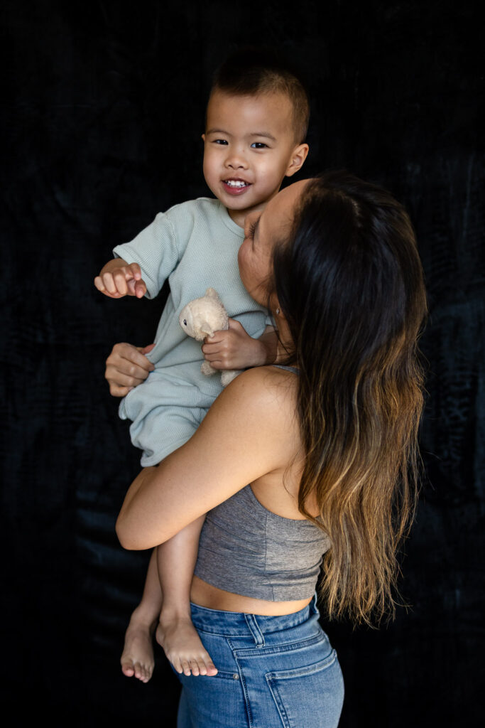 A mother holds her toddler on her hip while looking at him with affection as he smiles toward the camera during Mom and Me Photos