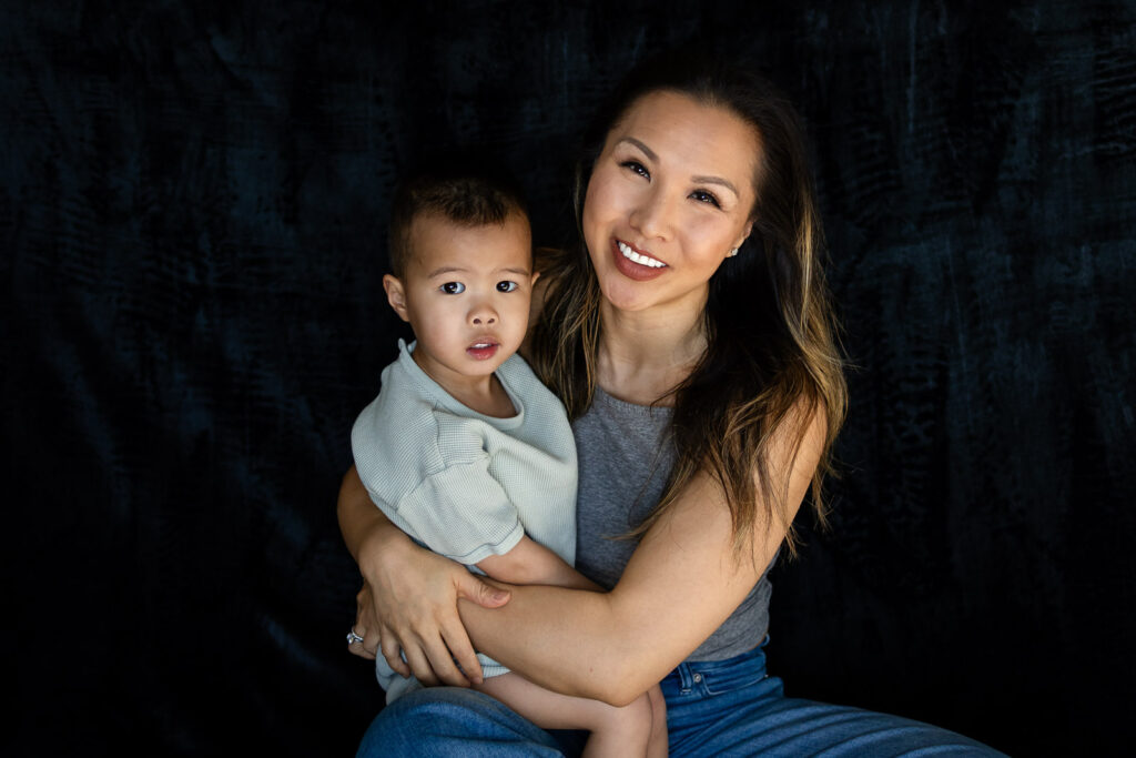A mother sits cross legged holding her toddler in her lap as they look at each other with calm loving expressions during Heartfelt Mama Minis