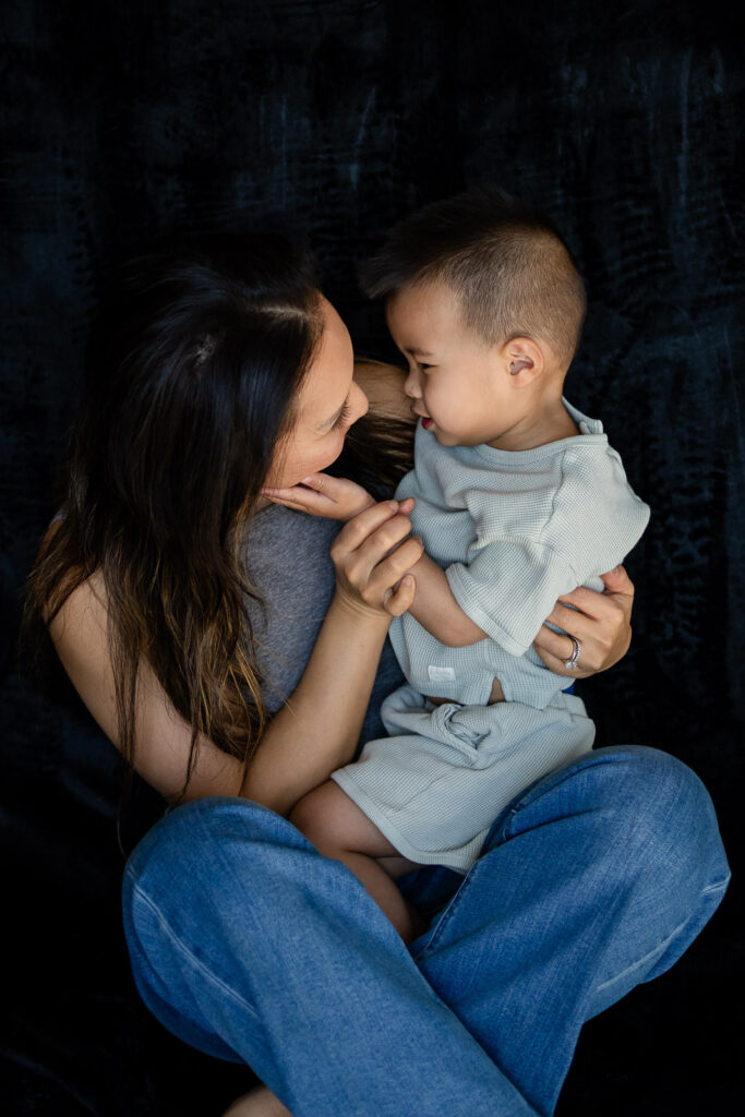 A mother sits cross legged holding her toddler in her lap as they look at each other with calm loving expressions during Mother & Child Photo Sessions