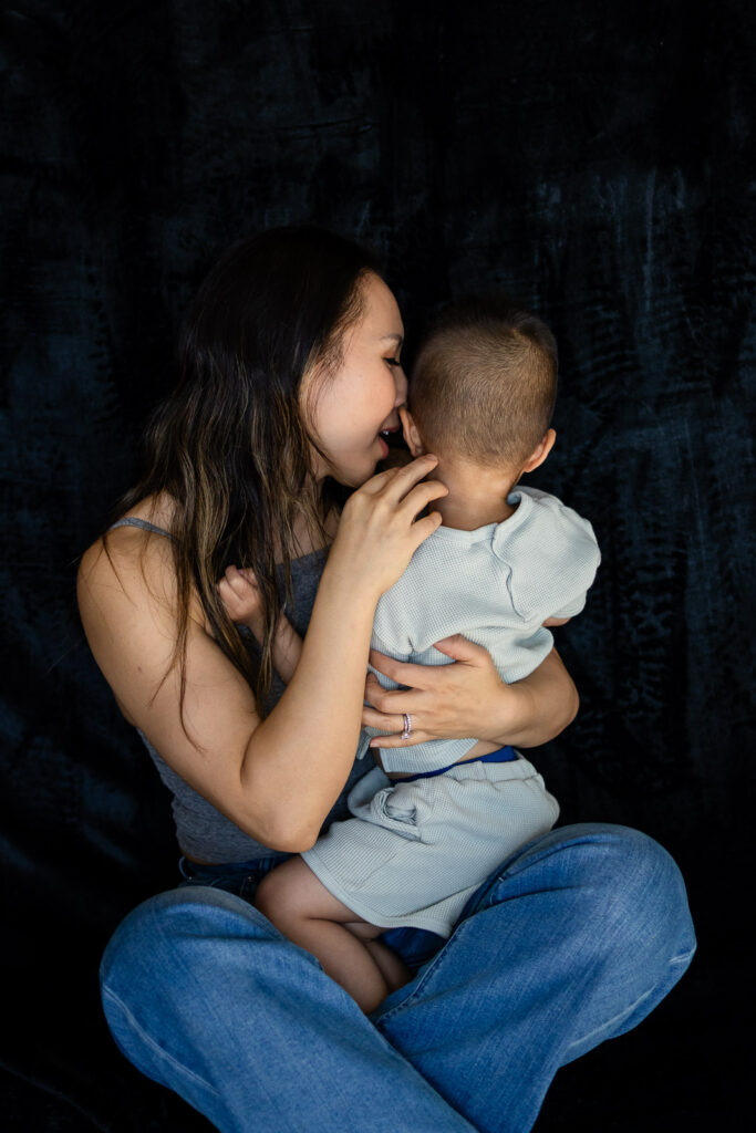 A mother kisses her toddler’s head as she cradles him close, creating a peaceful affectionate moment during Mother & Child Photo Sessions