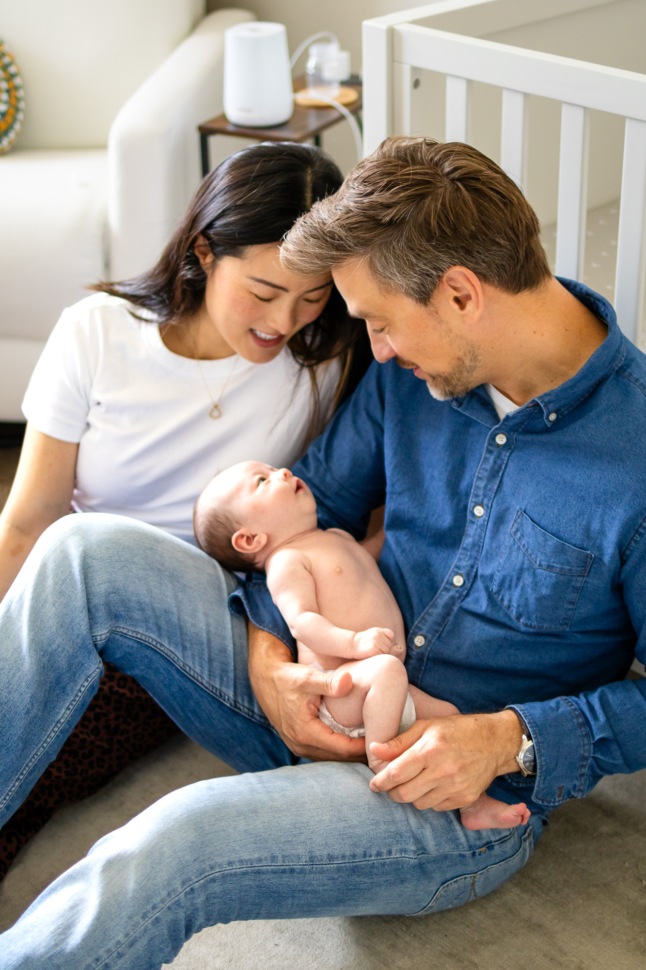 Parents sit close together holding their newborn near the crib during a peaceful in home newborn photography session.