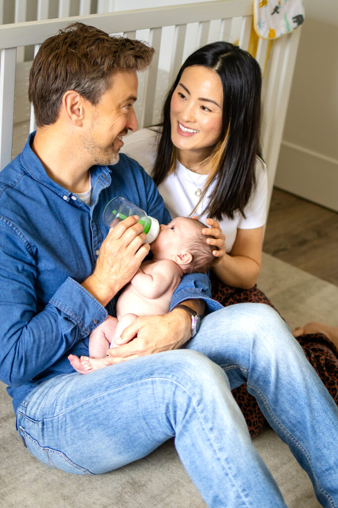A father feeds his newborn with a bottle while sitting on the nursery floor as the mother smiles and rests her hand on the baby’s head.