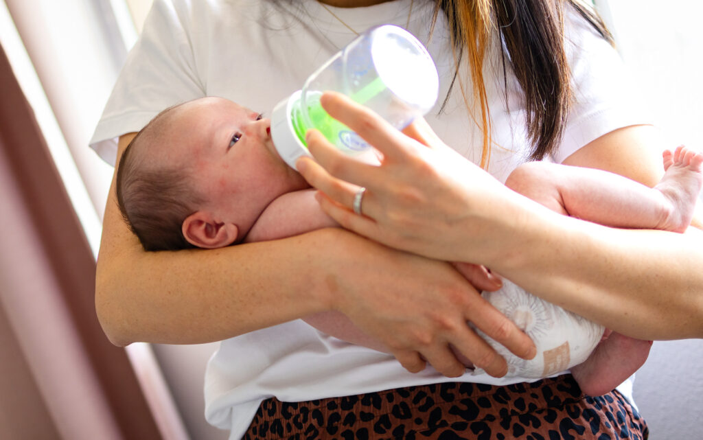 A mother bottle feeds her newborn while holding the baby securely in her arms in soft natural light.