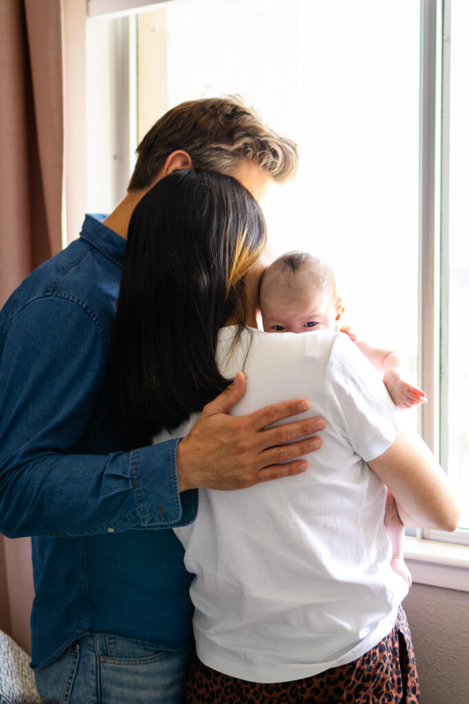 Both parents stand near a window holding their newborn together during newborn photos at home.