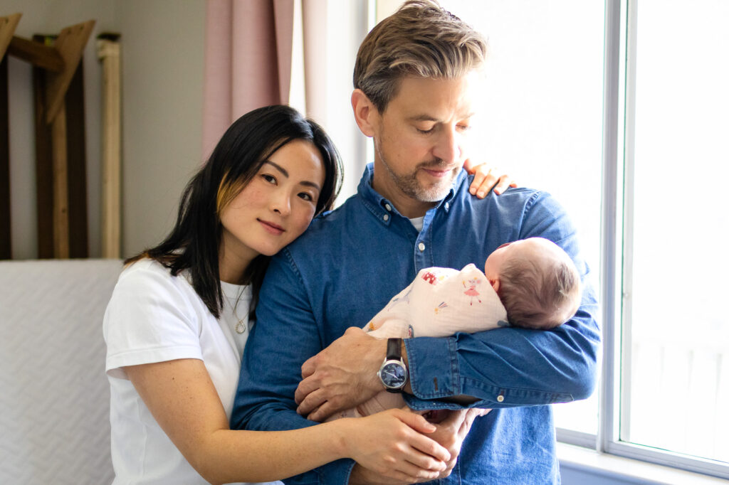 A mother leans her head on the father’s shoulder while he holds their sleeping newborn near the window.