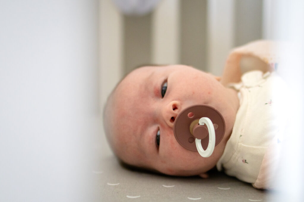 A newborn lies on a crib mattress with a pacifier while gazing softly toward the camera.