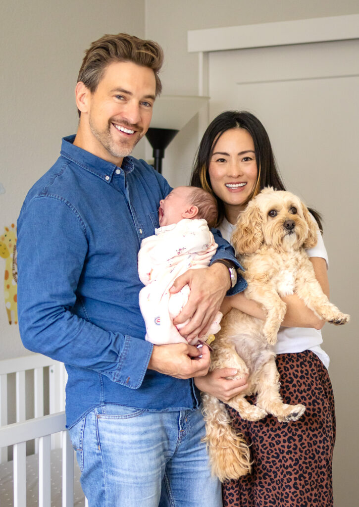 Parents stand in the nursery holding their newborn and family dog while smiling toward the camera.