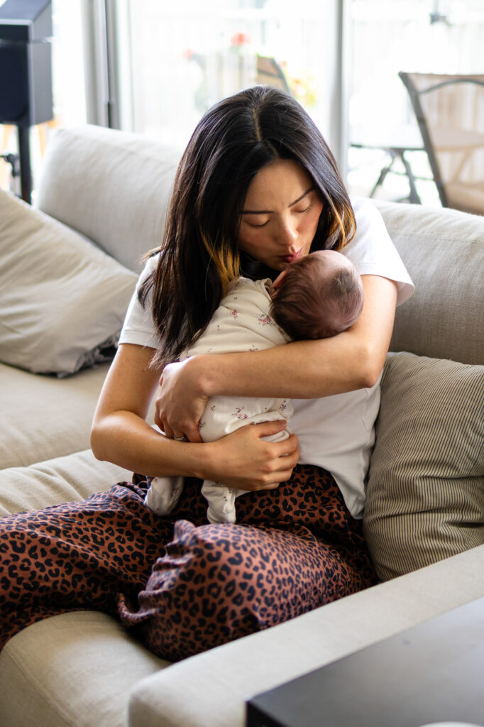A mother gently kisses her newborn while holding the baby in her arms during newborn photos at home in soft natural light.