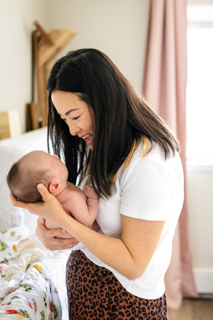 A mother smiles and talks to her newborn while holding the baby upright near the bed in their home.