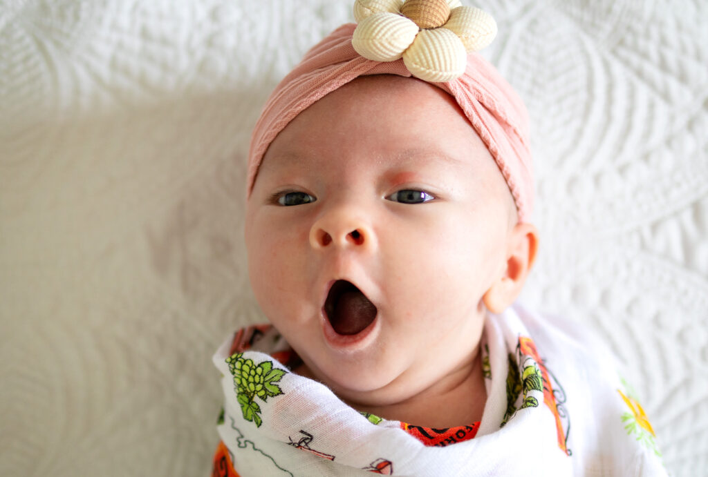 A newborn yawns while lying swaddled on a textured white blanket during newborn photos at home.