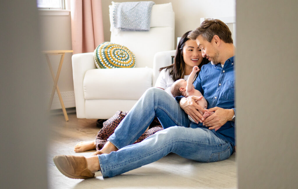 Both parents sit on the floor smiling down at their newborn as they share a gentle family connection.