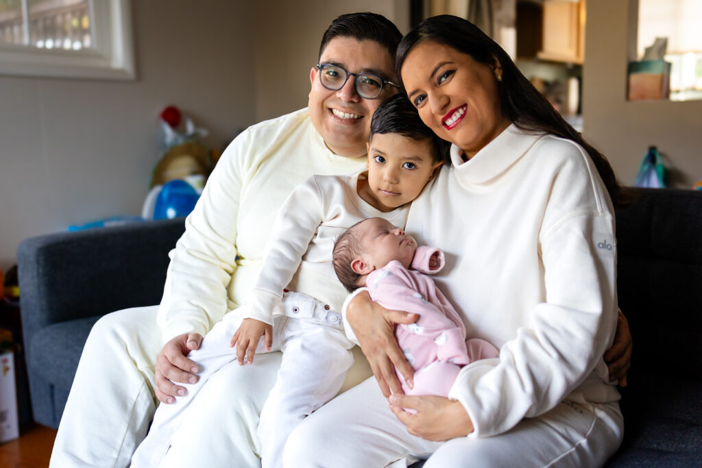 Parents sit close together on the couch while their older child leans in and a newborn sleeps peacefully in their arms, creating a cozy family portrait at home.