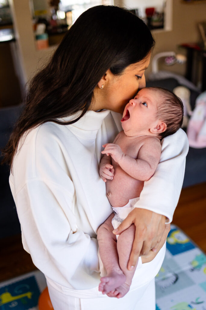 A mother gently kisses her newborn on the forehead while holding the baby close, capturing a quiet bonding moment at home.