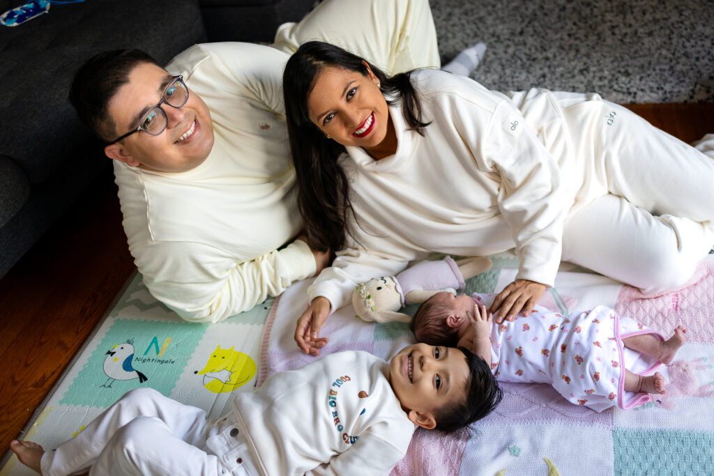 Parents and their older child lie close together on the floor while their newborn rests beside them, sharing a calm family moment.