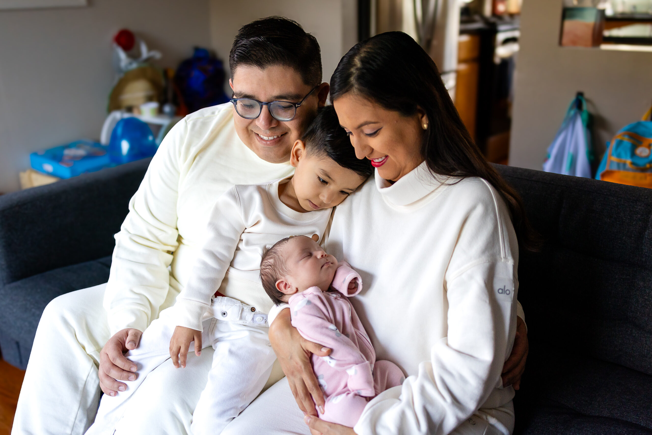 A mother and father look down lovingly as their older child gently rests beside them while they hold their sleeping newborn during a relaxed family moment.