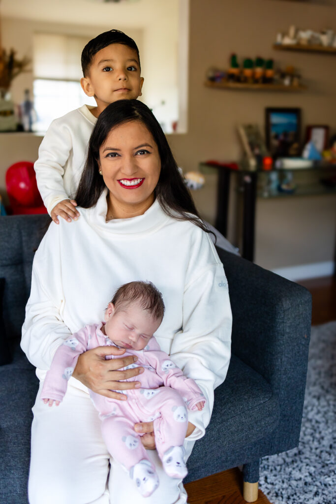 A mother smiles at the camera while holding her newborn, with an older sibling standing close behind her and resting their hands on her shoulders.