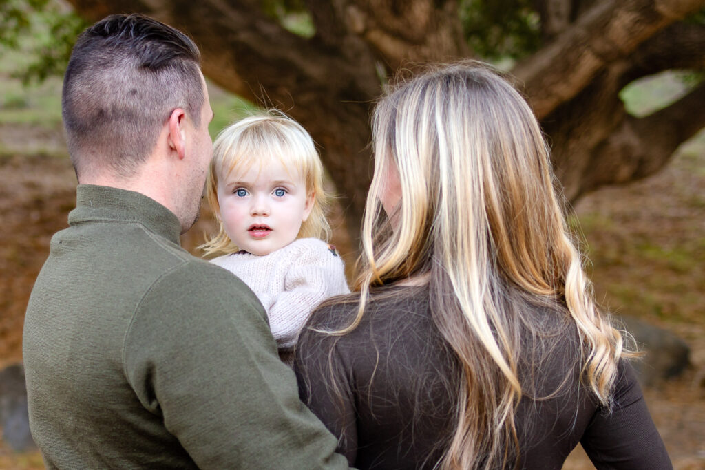 Parents stand close together holding their toddler between them while the child looks calmly toward the camera, framed by soft greenery in the park.