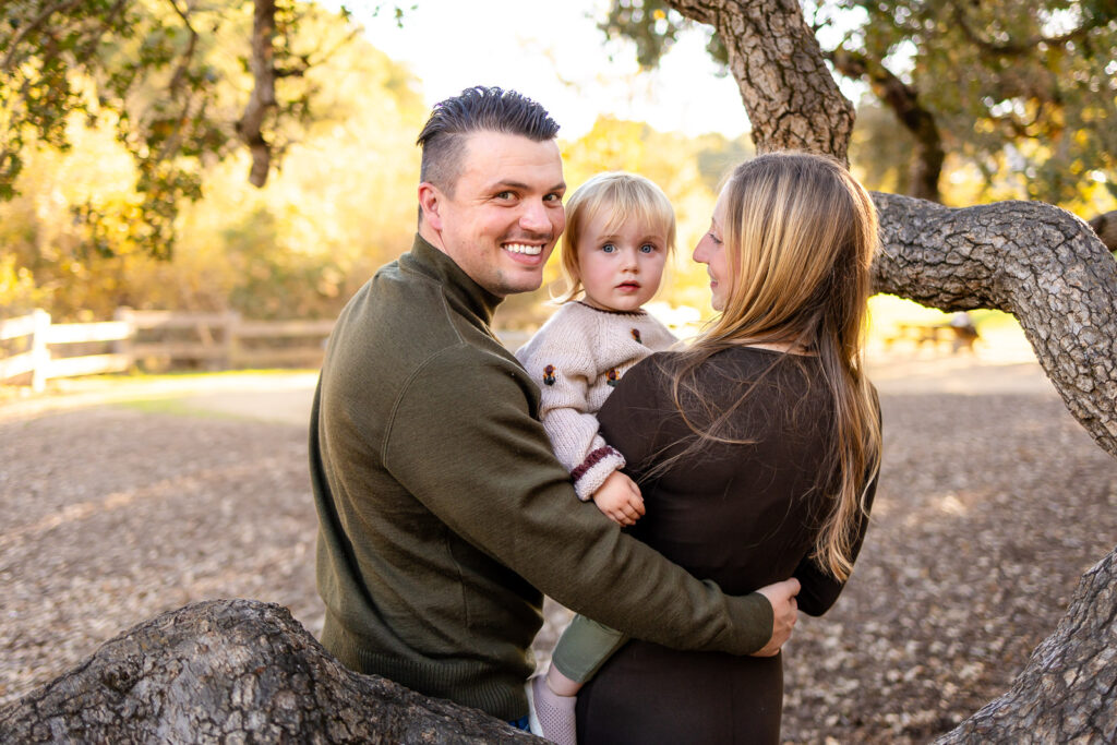 Parents hold their toddler and look toward the camera as the child gazes forward, surrounded by soft golden light and autumn leaves.