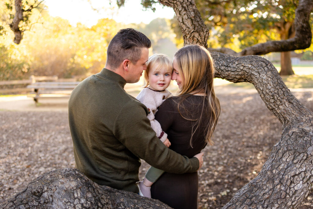 Parents lean in toward their toddler for a tender moment, sharing quiet smiles during an unhurried family photo session outdoors.