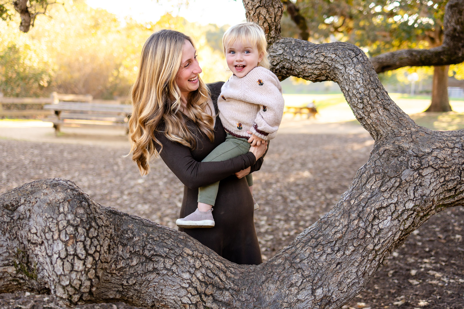 A mother holds her toddler and laughs as the child smiles brightly, standing within the curve of a tree branch at the park.