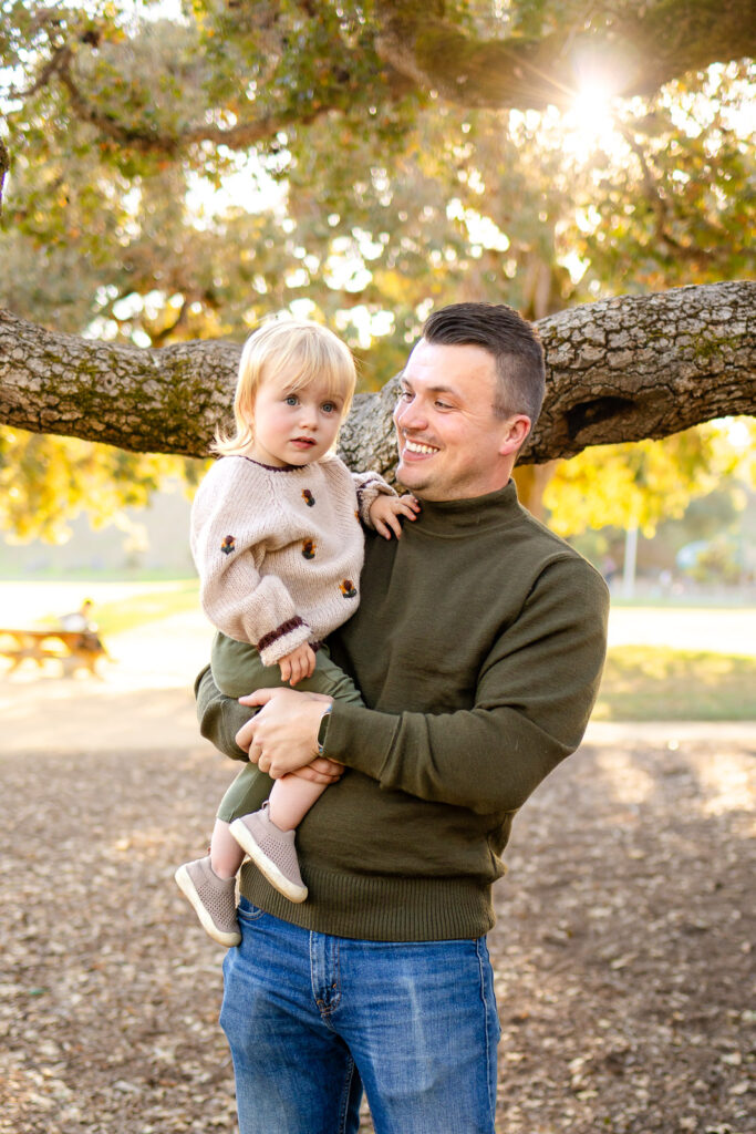 A father holds his toddler in his arms while smiling at them, capturing an easy and stress-free family photos moment under the trees.