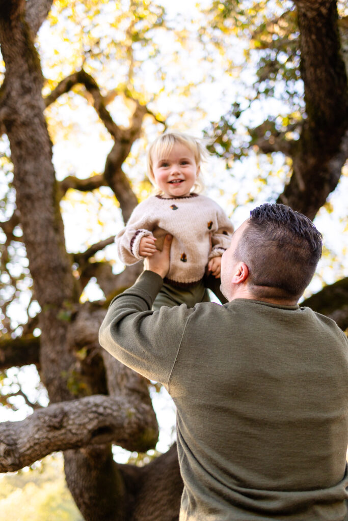 A father lifts his toddler high into the air as the child laughs, creating a playful and carefree family moment in nature.