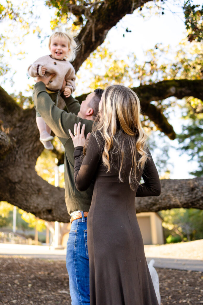 Both parents reach up toward their toddler as the child is lifted above them, sharing laughter and connection during a laid-back family shoot.