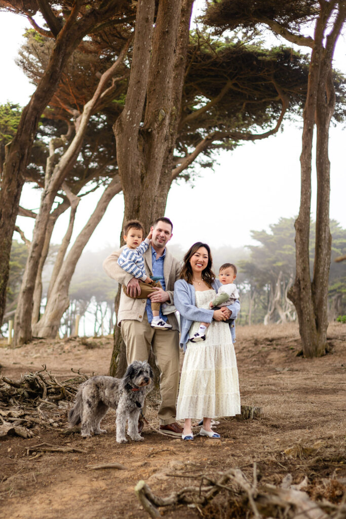 A family of four stands together in a quiet forest clearing with tall trees behind them while holding their two young children and their dog sits calmly at their feet.