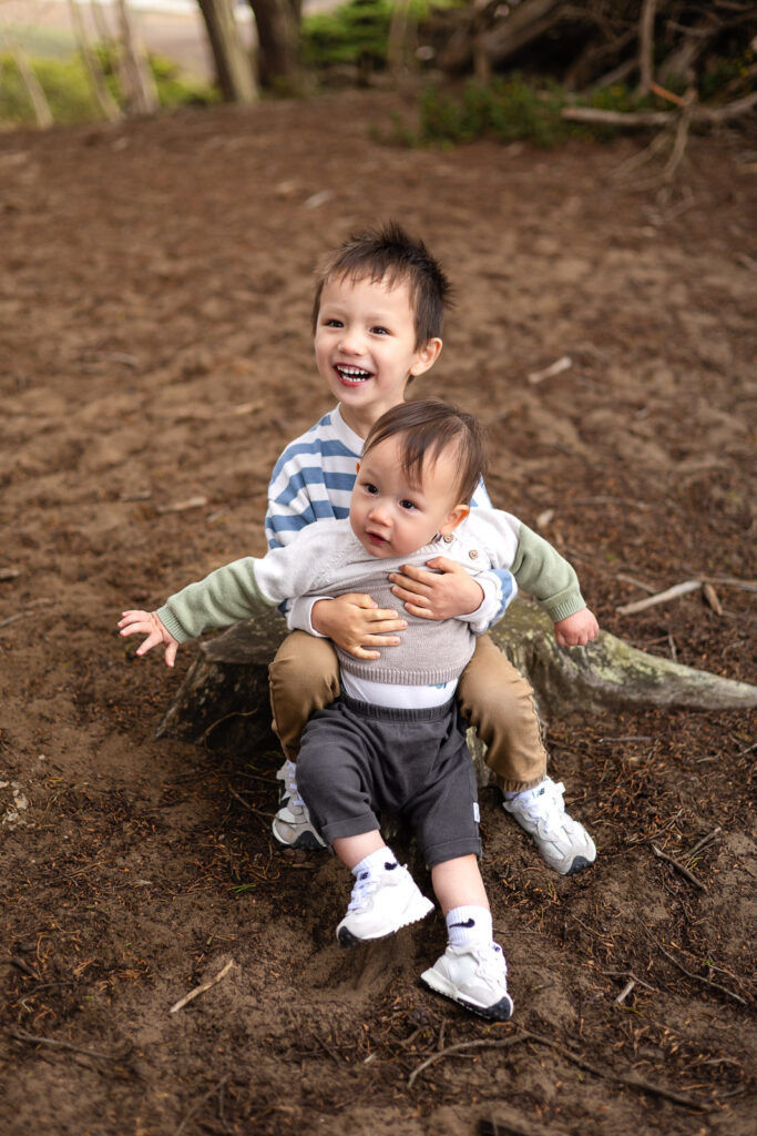Two young siblings sit together on a tree stump with one child hugging the other, showing a joyful and unposed sibling connection.