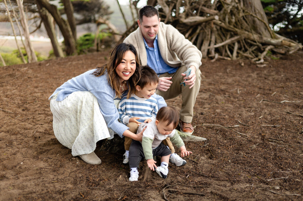 Parents crouch close behind their children as the toddler and baby explore the dirt together while being gently supported during a well led photo experience.