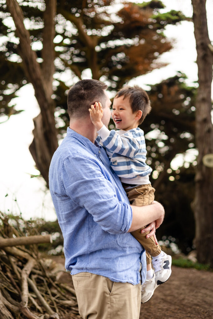 A father holds his toddler close as the child smiles and reaches for his face, sharing a tender and connected moment.
