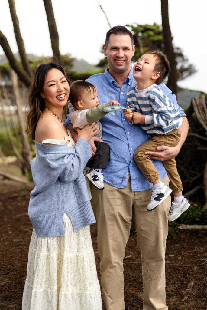 The family stands together beneath twisting tree branches with their dog at their side, posing comfortably during a well led photo experience.