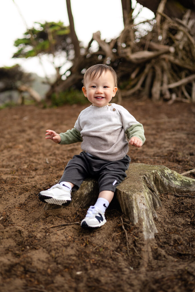 A baby sits on a tree stump smiling toward the camera while balanced comfortably in a calm outdoor setting.