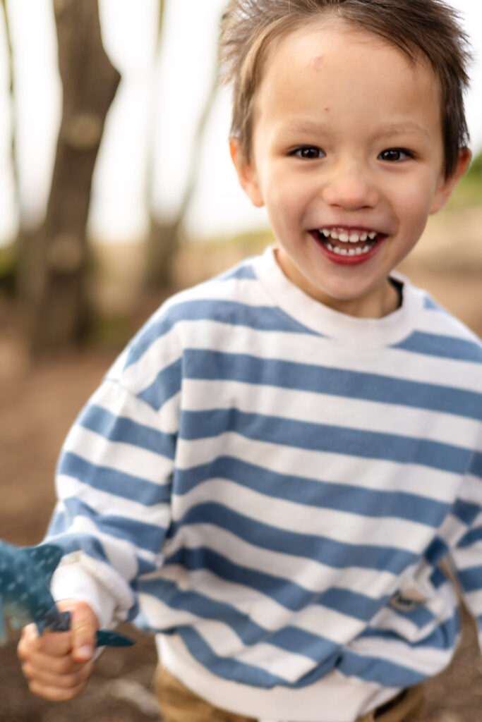 A close up portrait shows a toddler smiling brightly while holding a small toy, captured naturally in soft outdoor light.