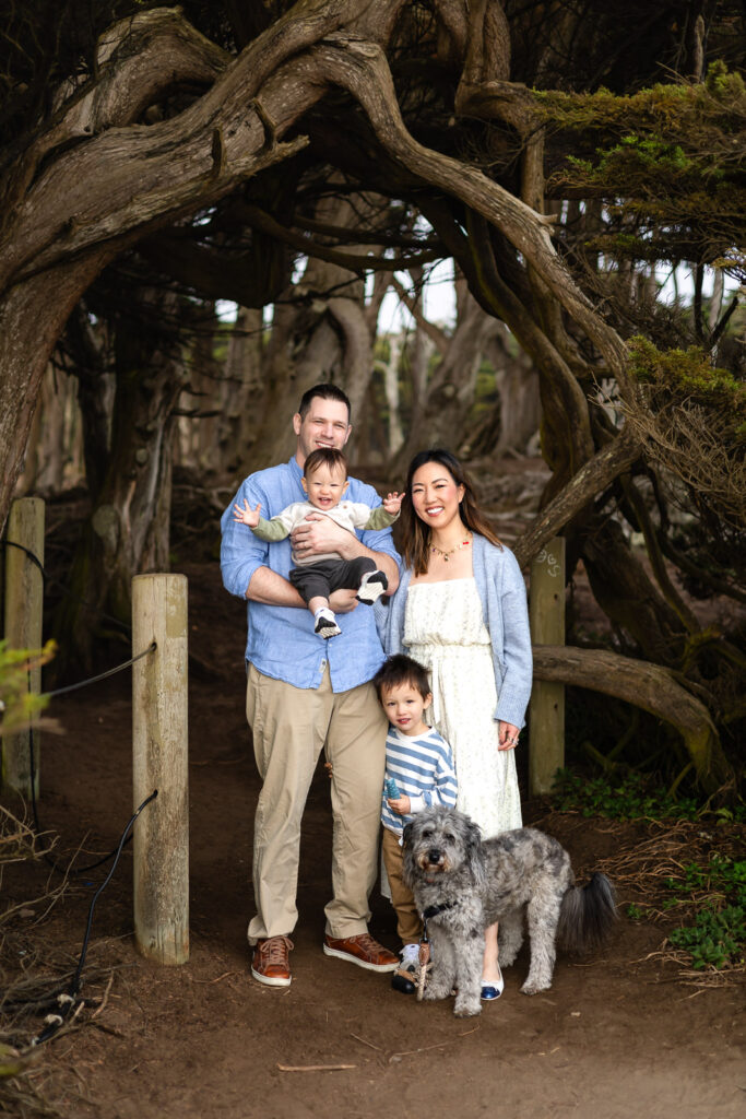 A family stands together beneath an arch of trees while holding their children and dog, creating a naturally guided and connected portrait.