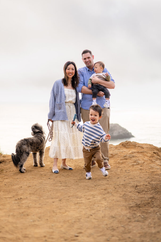 The family walks along a coastal overlook while the parents guide their children and dog with the ocean stretching behind them.