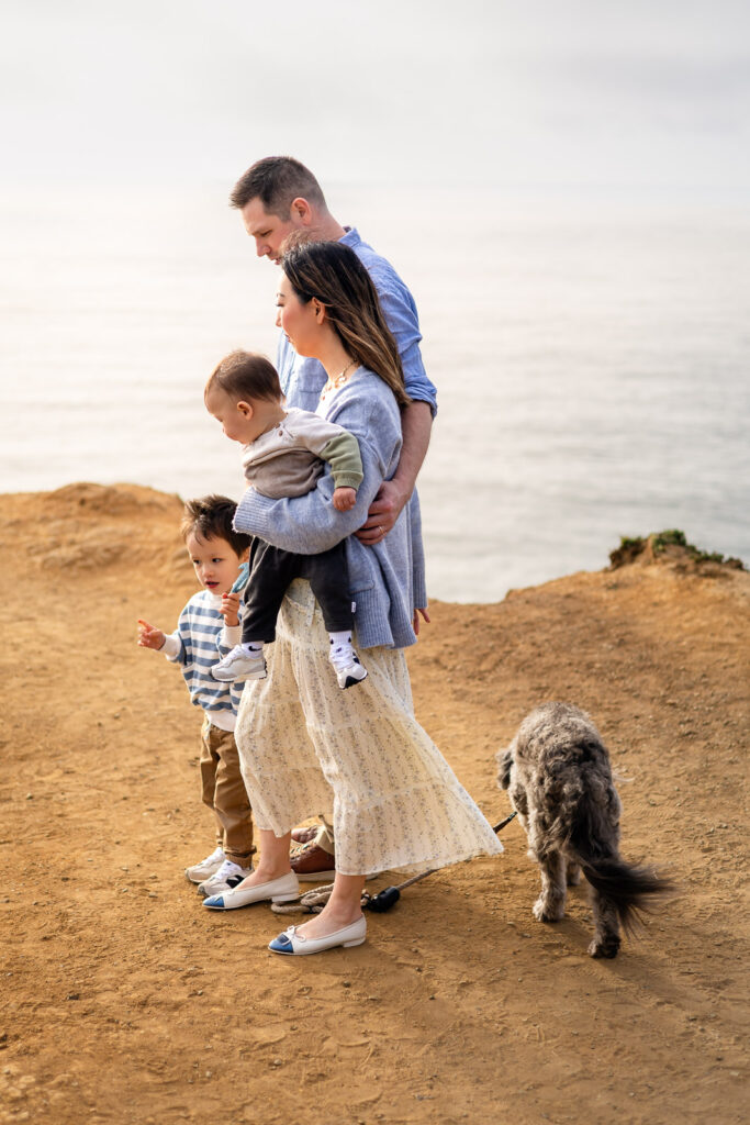 Parents stand close together holding their baby while the toddler walks ahead near the cliffside, showing a calm and supported family moment by the ocean.