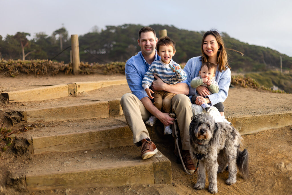 Parents sit on wooden steps with their two children close while their dog sits nearby, creating a relaxed and connected family moment.